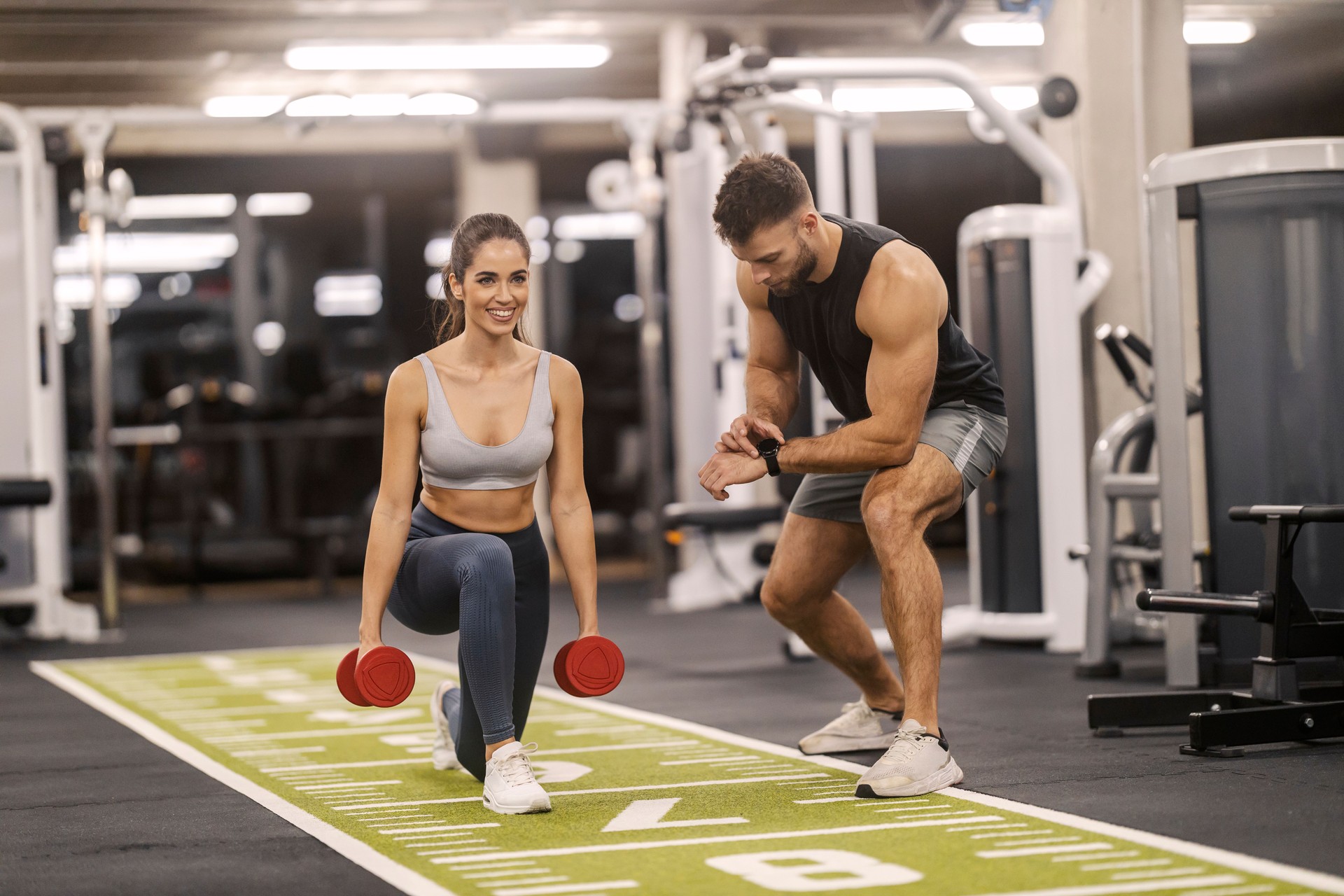Strong fit sportswoman doing lunges with dumbbells at gym with her trainer.